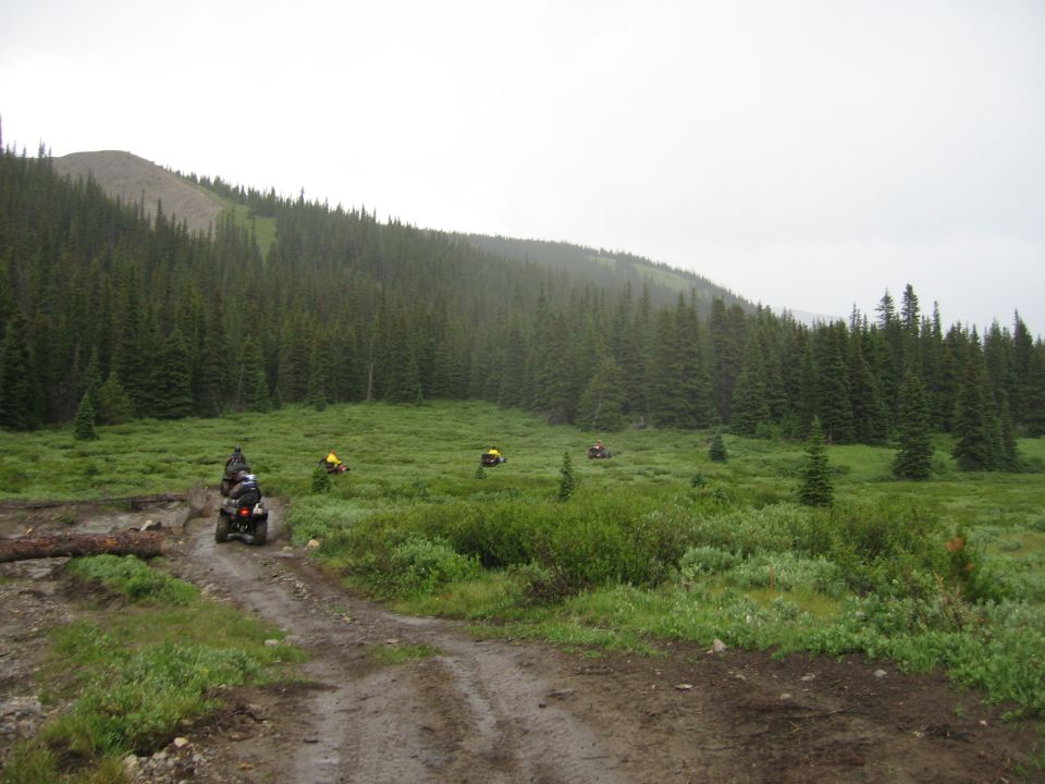 ATVs on a muddy trail with forest and mountain backdrop.
