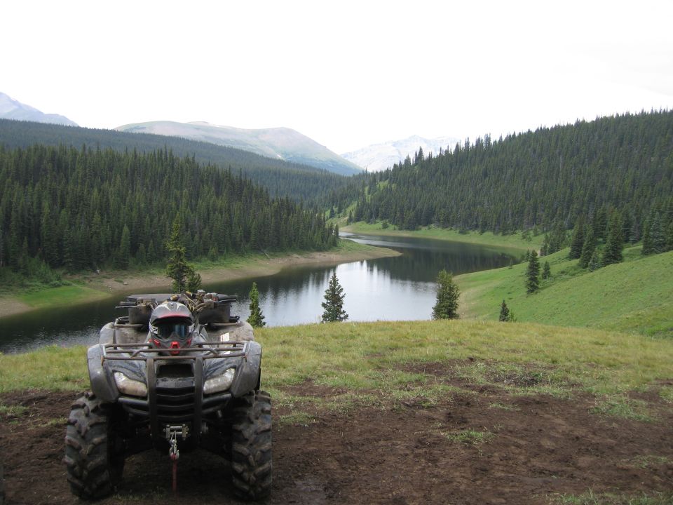 Dark grey ATV with helmet parked on a grassy hill overlooking a lake and forest.