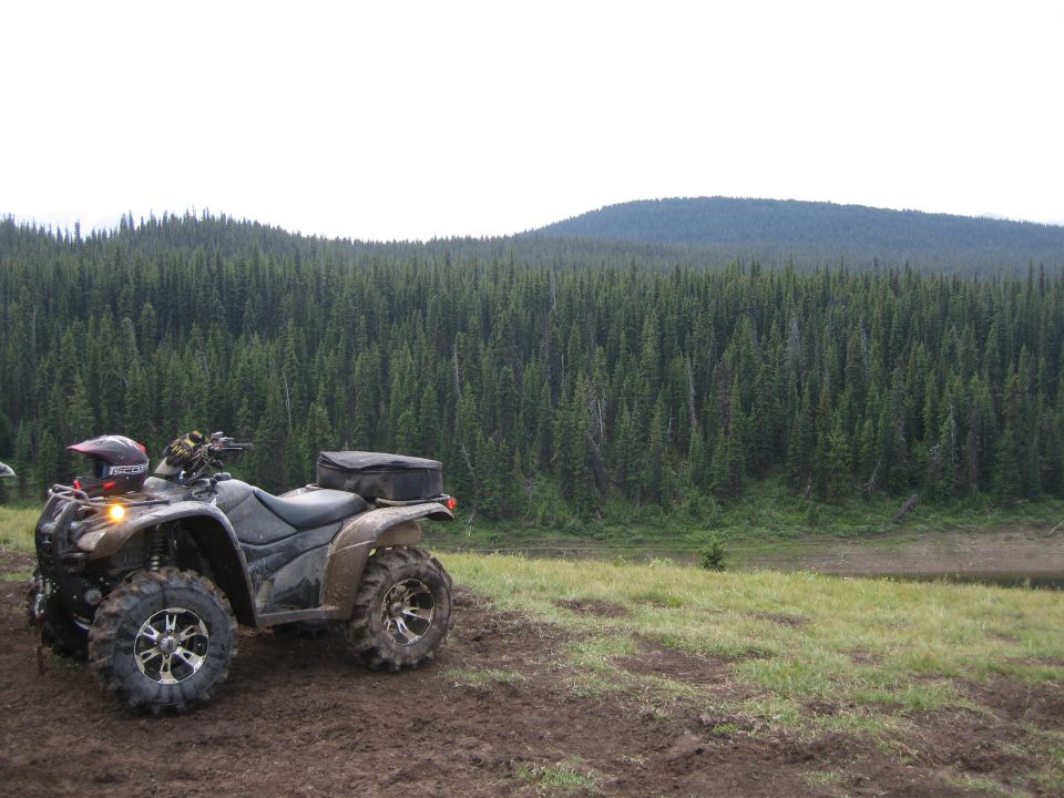 Black ATV parked on a muddy trail with a helmet on the handlebars, facing right, with a forest in the background.