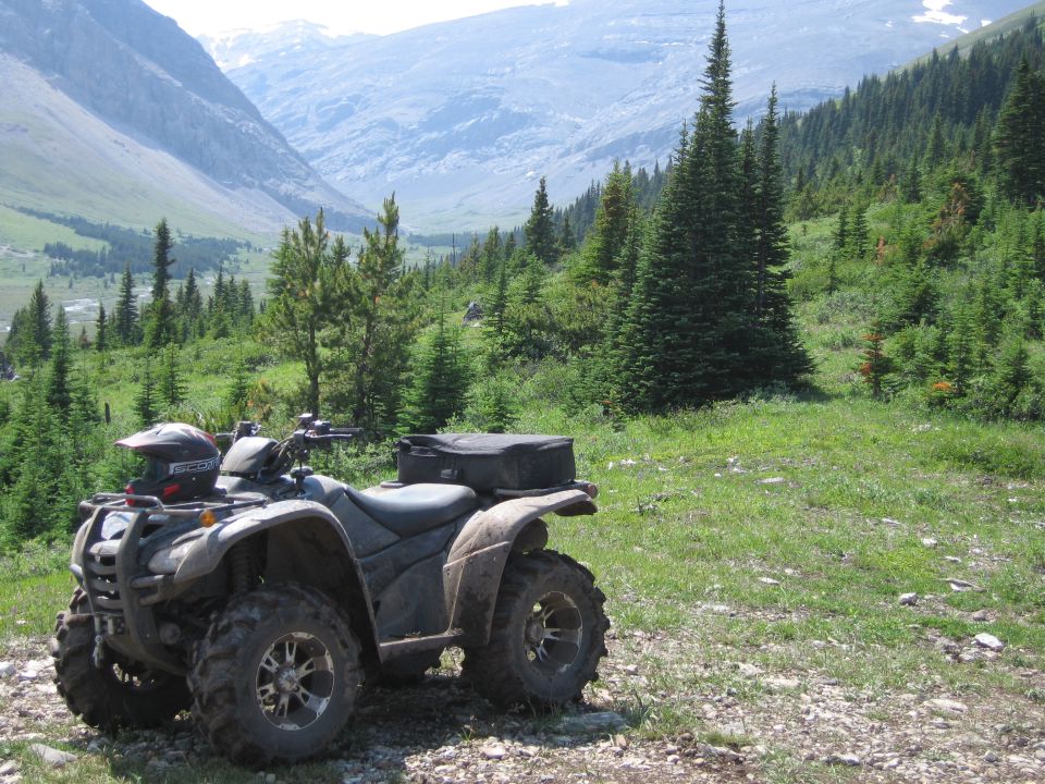 ATV parked on a gravel path with a helmet on the front rack, mountainous forest background.