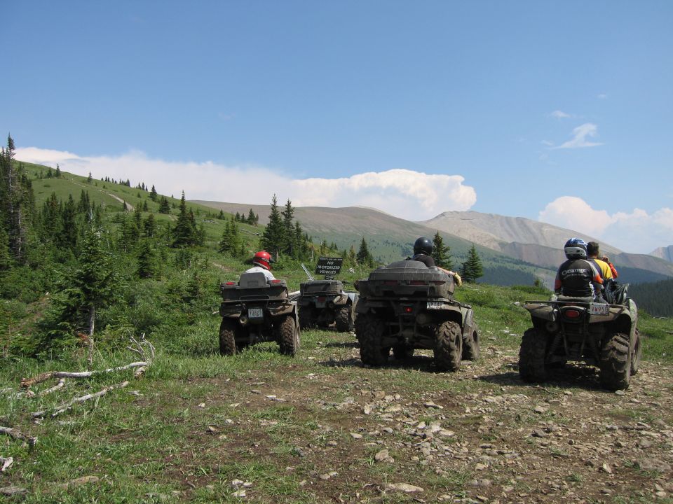 Four ATVs parked on a mountain trail with riders.