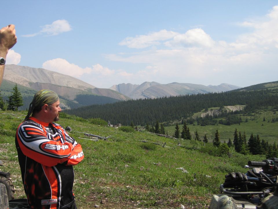 Man in orange and black jersey with arms crossed, overlooking a mountain and forest landscape. ATV in foreground.