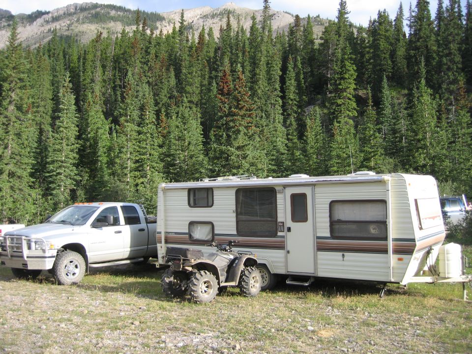 Silver pickup truck parked next to a travel trailer with an ATV hitched to the back, in a forest setting.