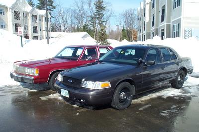 1991 Ford Crown Victoria LTD - Two sedans, a dark gray Ford Crown Victoria and a red older model, parked in a snowy lot.