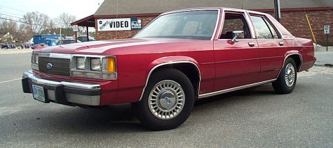 1991 Ford LTD Crown Victoria - Front three-quarter driver-side view of a burgundy Ford LTD Crown Victoria four-door sedan parked in front of a brick storefront with a "Vi…