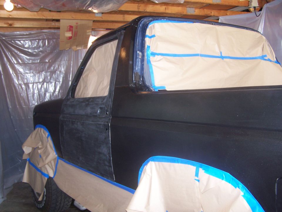 Side view of a dark-colored Ford Bronco, masked and taped for painting in a garage