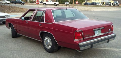 1991 Ford LTD Crown Victoria - A dark red Ford Crown Victoria LTD four-door sedan shot from the left rear three-quarter in a parking lot, with chrome window surrounds, bo…