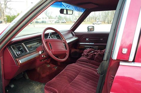 1991 Ford LTD Crown Victoria - Interior of a red boxy Ford full-size sedan shot through the open driver's door, showing a burgundy cloth bench seat, matching red dash and…