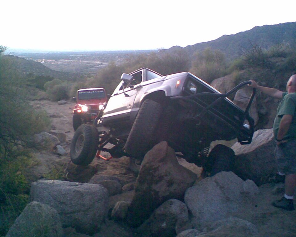 Off-road vehicle with roll cage navigating rocky terrain, red Jeep behind