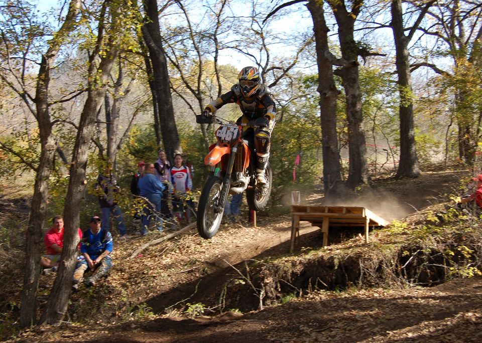 Orange KTM dirt bike jumping over wooden ramp on wooded trail with spectators