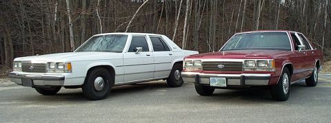 1991 Ford LTD Crown Victoria - Two sedans, a white and a red one, are parked side-by-side. The red sedan features a grille characteristic of 1980s Ford models.