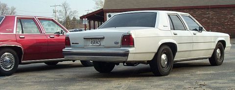 1991 Ford LTD Crown Victoria - Rear three-quarter view of a white Ford LTD Crown Victoria sedan on black steel wheels with no hubcaps, wearing a Massachusetts plate readi…