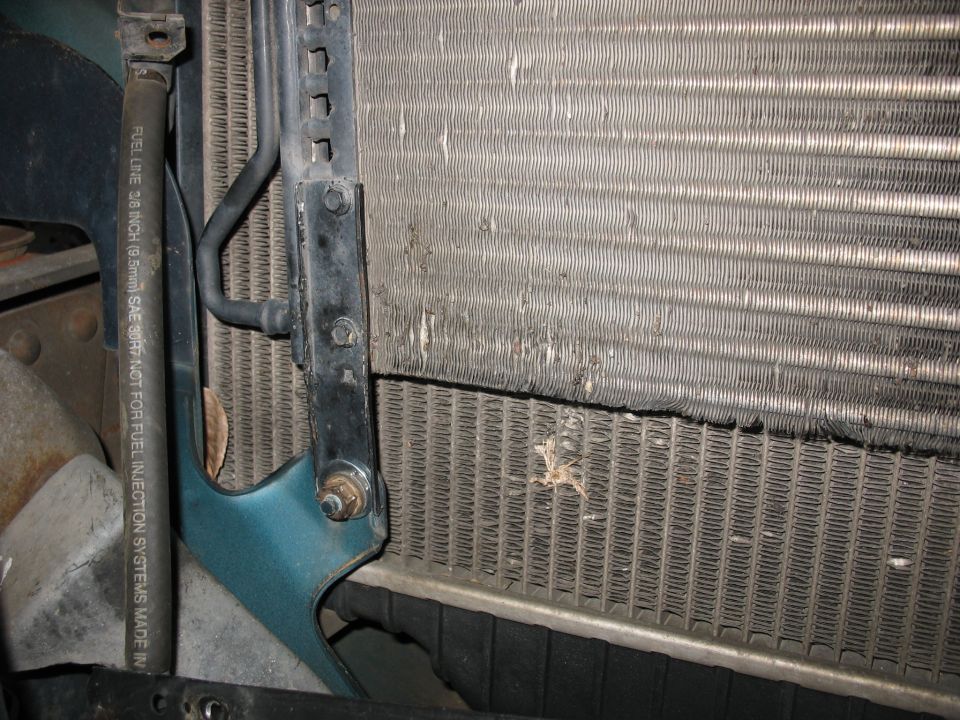 Close-up of a damaged radiator and intercooler assembly with visible hoses and brackets. A fuel line has text printed on it