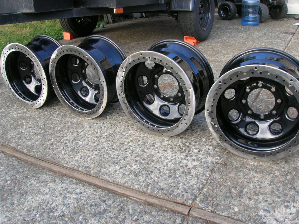Four black off-road wheels with polished metal lips sit on a concrete surface next to a trailer