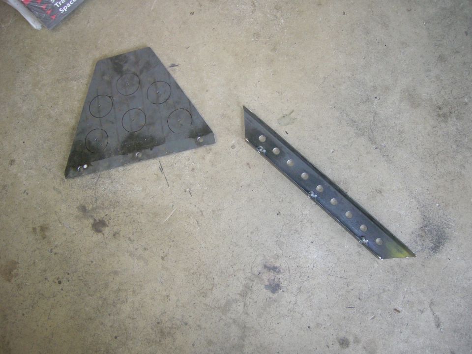 Two metal pieces, one triangular with circles drawn on it, the other a long perforated strip, lie on a concrete floor
