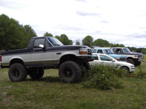 1993 Ford Bronco - A heavily modified pickup truck with oversized tires is parked in a grassy field alongside other vehicles.