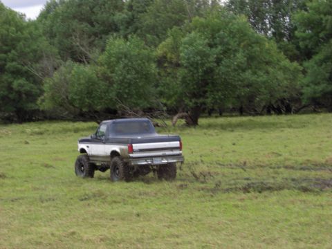 1993 Ford Bronco - Lifted pickup truck navigates a grassy, muddy field with trees in the background.