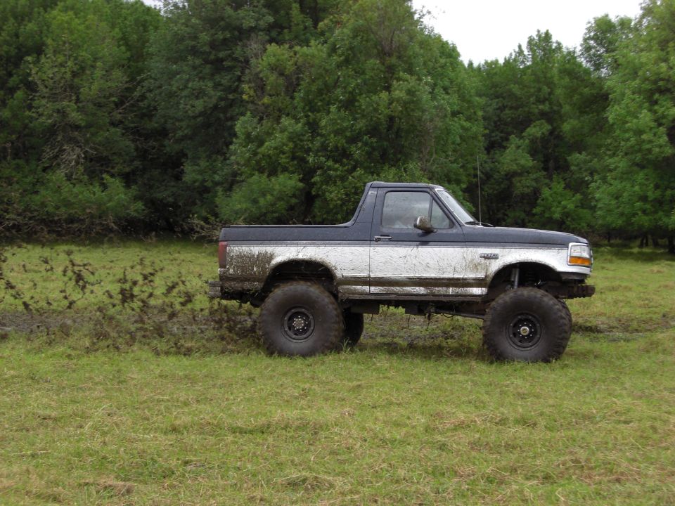 Lifted pickup truck with large tires driving through mud and grass, trees in background