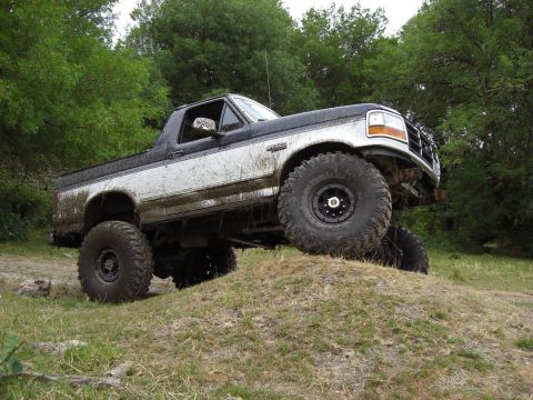 1993 Ford Bronco - A lifted, mud-covered Ford Bronco navigates an off-road obstacle on a grassy incline.