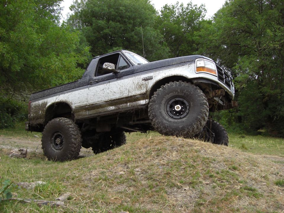 Mud-splattered two-tone Ford Bronco climbing a grassy hill, front right wheel elevated