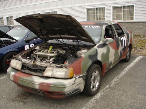 1992 Ford Taurus - A 1992 Ford Taurus, painted in camouflage, sits with its hood open at a convention.