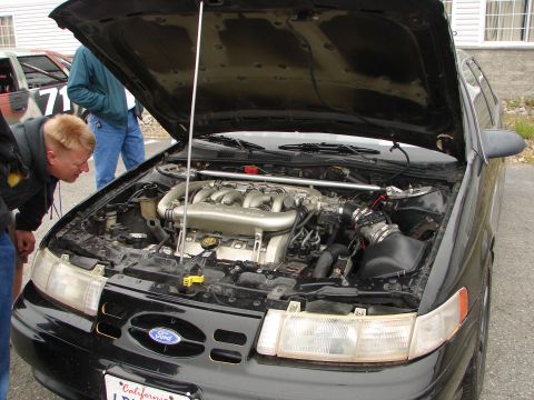 1992 Ford Taurus - A black Ford Taurus's engine bay is displayed, with onlookers examining the motor. Another vehicle is partially visible in the background.