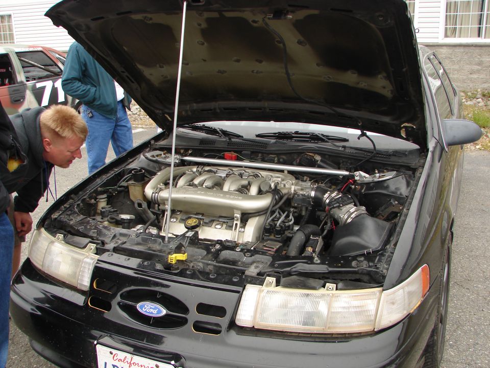 Black Ford Taurus with hood open, showing engine. People looking at engine. Another car with number 71 visible in background