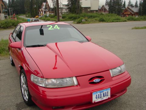 1992 Ford Taurus - A red 1992 Ford Taurus is parked outside with houses and trees in the background.