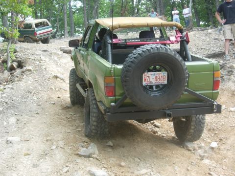 1995 Ford Bronco - Two SUVs navigate a rocky off-road trail. The foreground vehicle is green with a spare tire on the back.