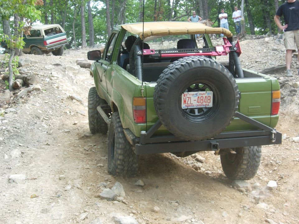 Green SUV on rocky trail, spare tire mounted on rear bumper. Another SUV is visible in the background