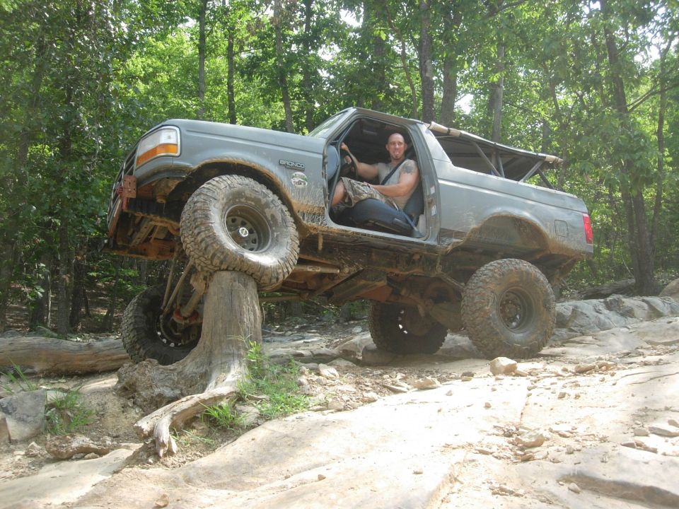 Man driving a lifted Ford Bronco on a tree stump off-road.