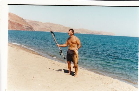 1994 Ford Bronco - A man stands on a beach holding a speargun and two fish, with the ocean and hills behind him.