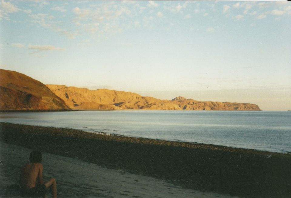 Person sitting on a beach facing the ocean and rocky hills