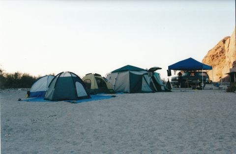 1994 Ford Bronco - A campsite scene features several tents set up on a sandy beach, with a dark SUV parked under a blue canopy near a cliff face.