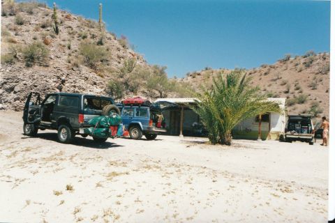 1994 Ford Bronco - Several vehicles are parked in a sandy, desert area near a small building, with a person standing by one of the vehicles.
