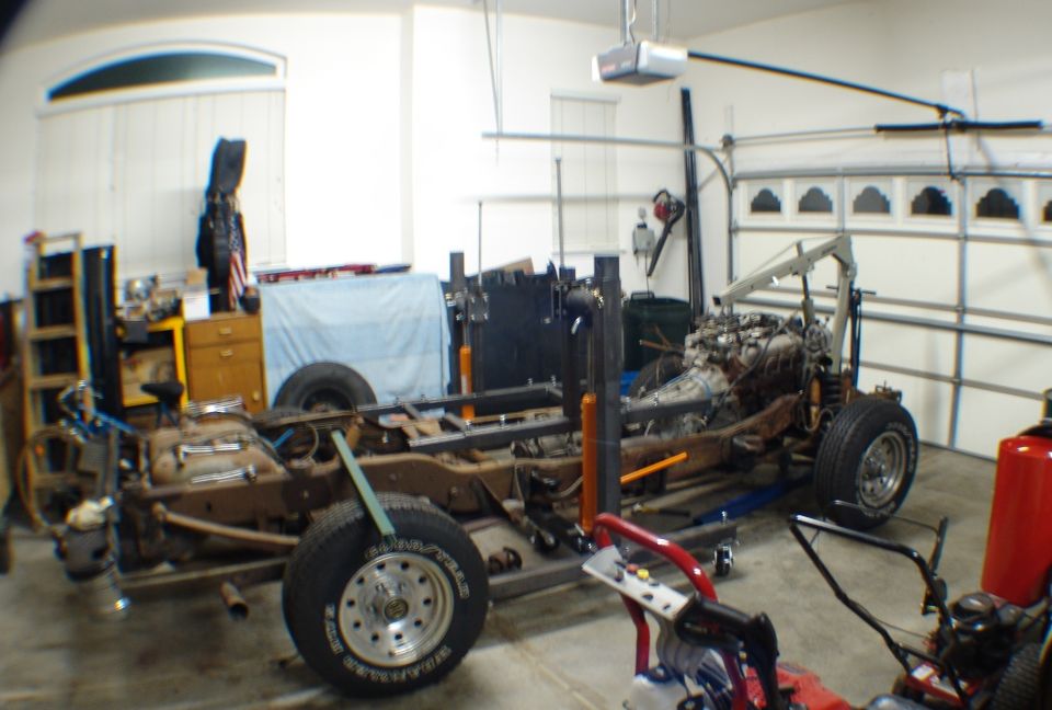 Stripped truck chassis with engine and transmission on stands in a garage next to an engine hoist