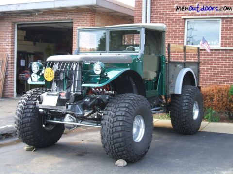 1991 Ford Bronco - This heavily modified truck sits on massive off-road tires, its green body contrasting with the brick facade behind it.