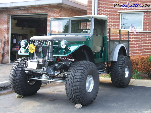 Green modified truck with oversized tires and a canvas top, parked in front of a brick building