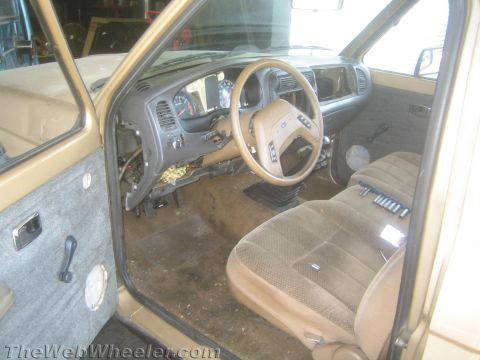 1990 Ford Bronco - tan interior of a Ford truck shows a cloth bench seat, steering wheel and partially stripped dashboard, viewed through the open door.