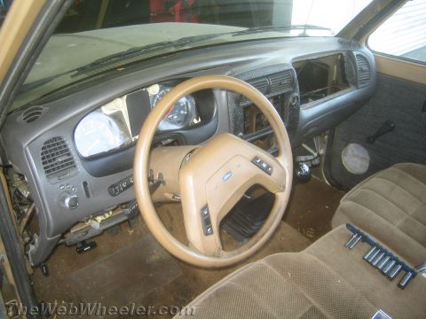 1990 Ford Bronco - Close interior view of a tan Ford truck showing the steering wheel, instrument cluster and cloth seat through the open driver door.