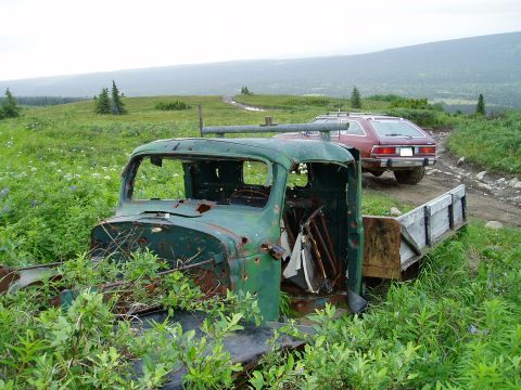 2000 Ford E350 - An abandoned green truck sits in a field, with a red wagon driving away on a dirt path in the distance.