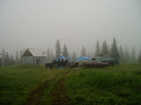 2000 Ford E350 - A collection of vehicles are parked in a clearing next to a small cabin, surrounded by trees and shrouded in fog.