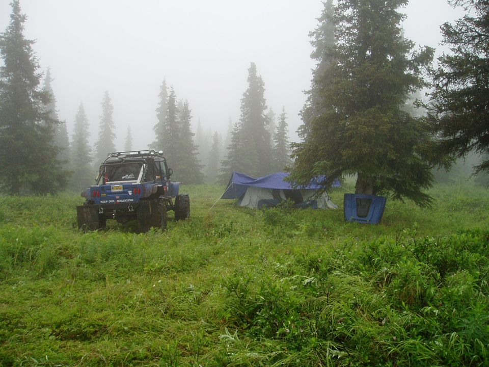 Blue off-road vehicle parked in a grassy field with tents and trees in the fog