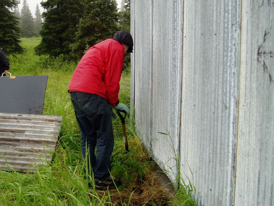 Person in red jacket digging near corrugated metal wall