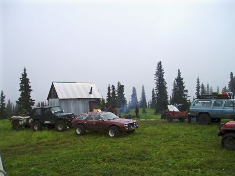 2000 Ford E350 - A group of off-road vehicles are gathered in a field next to a cabin, with trees and a cloudy sky overhead.