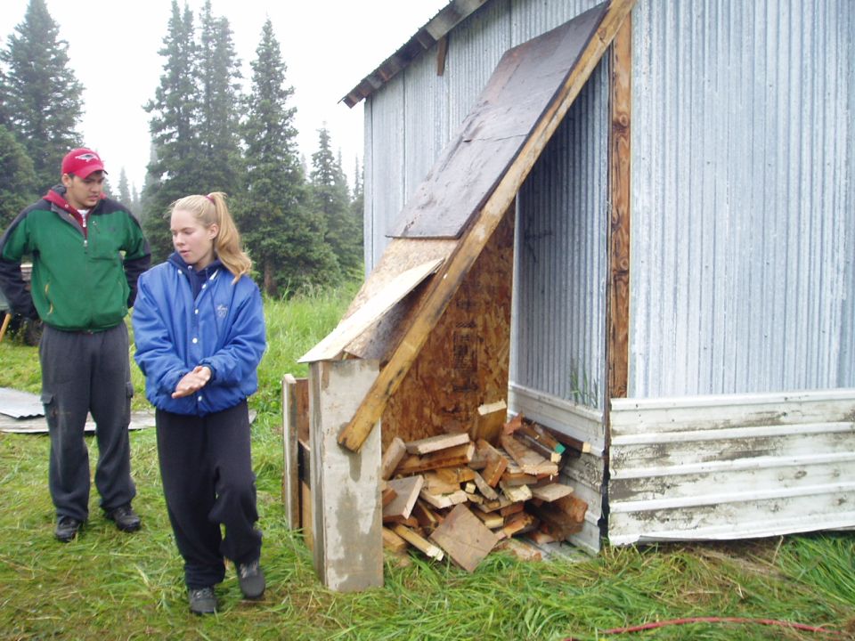 Two people stand near a shed with firewood stacked inside. A "SuperMotors.net" logo is visible in the lower right corner