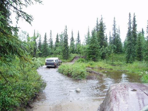 2000 Ford E350 - A dark pickup truck with a white camper shell navigates a muddy, water-filled trail surrounded by dense forest.