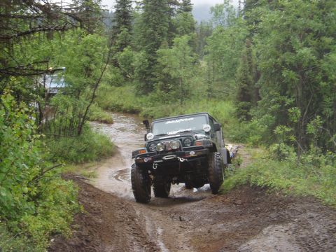 2000 Ford E350 - A dark green Jeep Wrangler is driving on a muddy, wooded trail, crossing a shallow stream. The Jeep has large tires, auxiliary lights, and…
