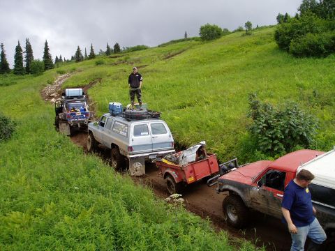 2000 Ford E350 - A convoy of off-road vehicles, including a lifted blue SUV towing a trailer, navigates a muddy, grassy hillside trail under an overcast sky…