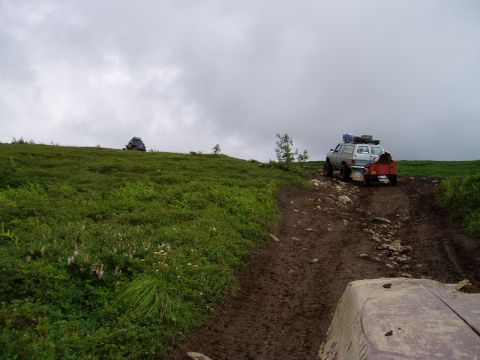 2000 Ford E350 - Two off-road vehicles are on a muddy trail on a grassy hillside under a cloudy sky. One vehicle, a light-colored SUV with a trailer, is asc…
